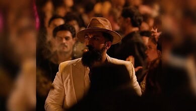 Man with a beard and hat in a crowded, dimly lit scene from a film or event.
