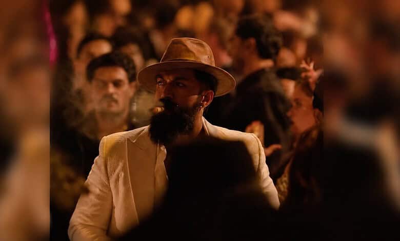 Man with a beard and hat in a crowded, dimly lit scene from a film or event.