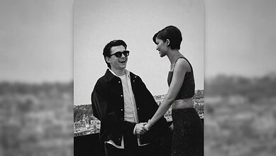 A smiling man and woman holding hands outdoors, with a cityscape in the background.