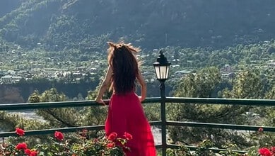Woman in a red dress overlooking a scenic valley with mountains and greenery.