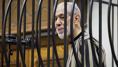 A man with a grey beard and striped shirt behind bars in a courtroom.