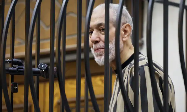 A man with a grey beard and striped shirt behind bars in a courtroom.