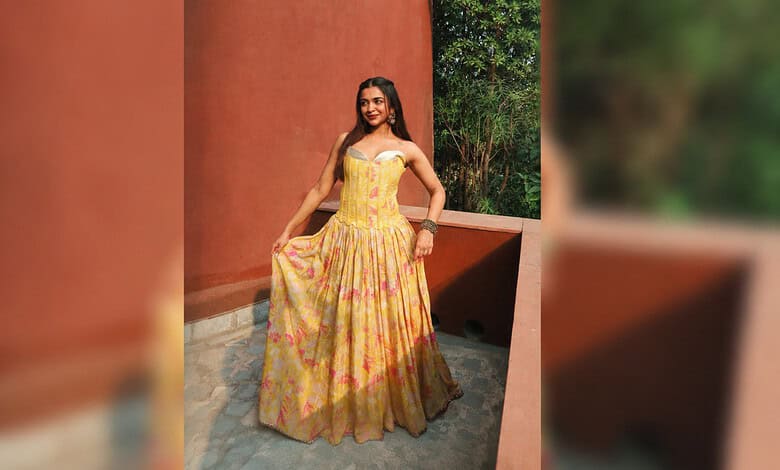 A woman in a yellow floral dress standing outdoors against a red wall and green foliage.