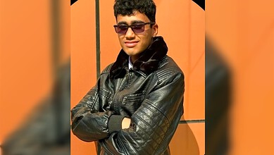 Young man wearing sunglasses and a leather jacket with arms crossed, smiling confidently.
