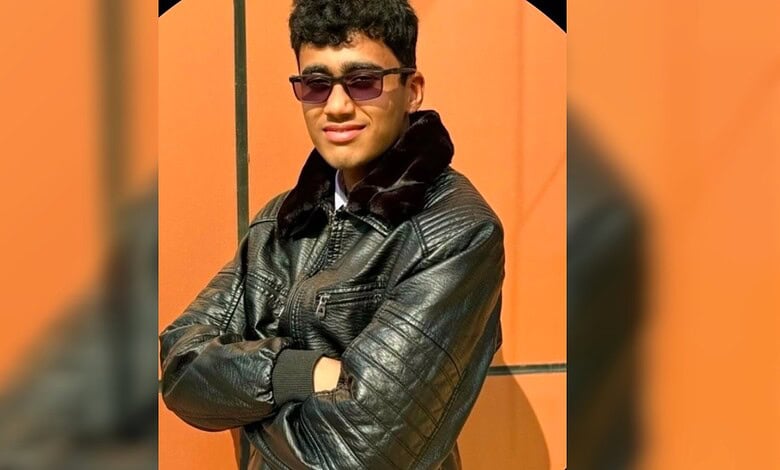 Young man wearing sunglasses and a leather jacket with arms crossed, smiling confidently.