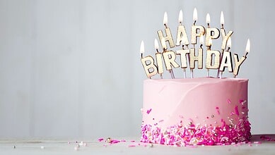 Pink birthday cake with lit candles spelling 'HAPPY BIRTHDAY' and pink sprinkles.