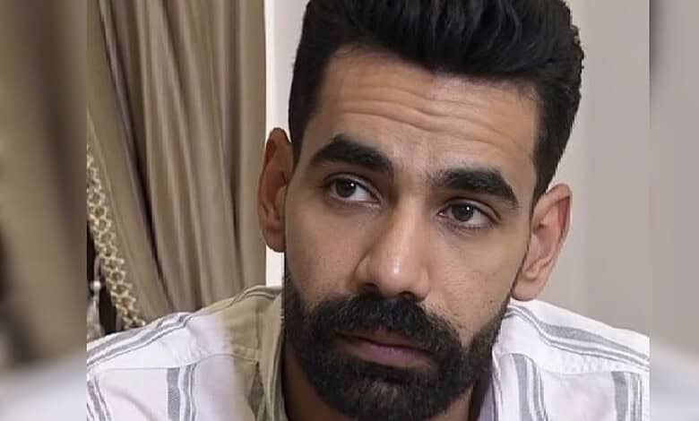 Close-up of a man with dark hair and beard wearing a white shirt, looking thoughtful.