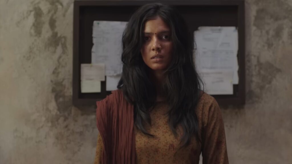 Malvika Mohanan in a brown outfit with long black hair, looking serious, in front of a notice board.