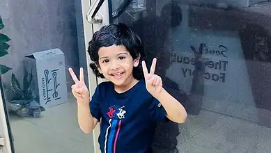 Smiling toddler making peace signs with both hands, wearing a navy T-shirt, indoors near a glass door.