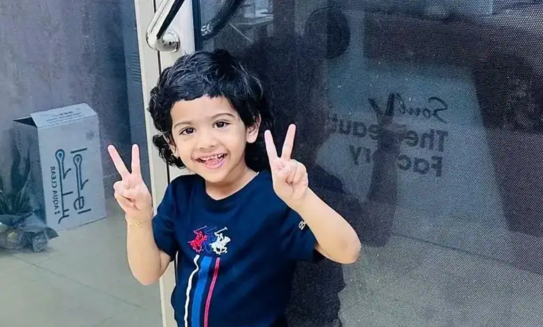 Smiling toddler making peace signs with both hands, wearing a navy T-shirt, indoors near a glass door.