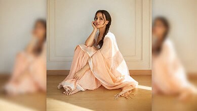 Elli AvrRam sitting on the floor in a light peach dress, smiling softly, in a minimalistic room.
