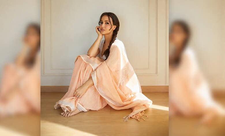 Elli AvrRam sitting on the floor in a light peach dress, smiling softly, in a minimalistic room.