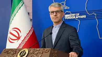 Iran's Foreign Ministry spokesperson speaking at a press conference with Iran flag in background.