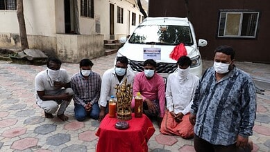 Six men with a Panchaloha idol and bottles, seated on the ground in front of a white vehicle, wearing fac.