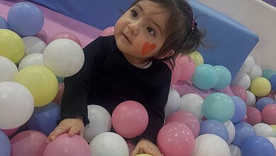 Adorable girl with face paint in a colourful ball pit, looking up with curiosity and wonder.