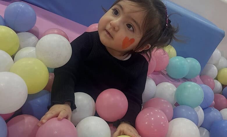 Adorable girl with face paint in a colourful ball pit, looking up with curiosity and wonder.