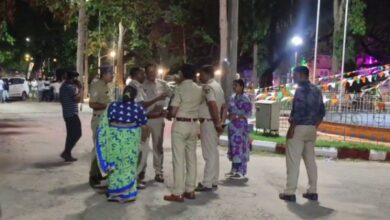 Police officers speaking with two women on a street at night, with tricolor bunting decorating a park in the background.