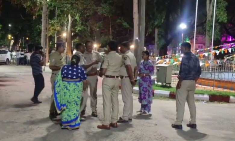 Police officers speaking with two women on a street at night, with tricolor bunting decorating a park in the background.