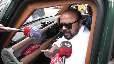 Man speaking to media with microphones inside a vehicle, wearing sunglasses and white shirt.