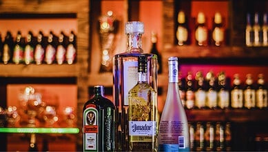 Bottles of Jägermeister, Absolut vodka, El Jimador tequila, and Hpnotiq liqueur lined up on a bar counter with blurred shelves of bottles in the background.