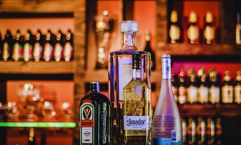 Bottles of Jägermeister, Absolut vodka, El Jimador tequila, and Hpnotiq liqueur lined up on a bar counter with blurred shelves of bottles in the background.