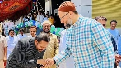 Political leaders shaking hands at a public event with a crowd in the background.