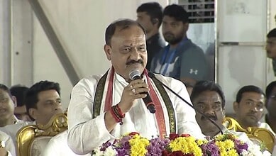 Indian politician speaking at a public event with a microphone, surrounded by audience members.