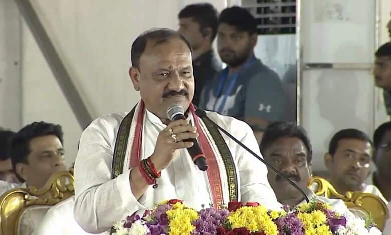 Indian politician speaking at a public event with a microphone, surrounded by audience members.