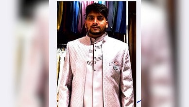Young man wearing a cream-coloured traditional outfit in a clothing shop with colourful garments behind.