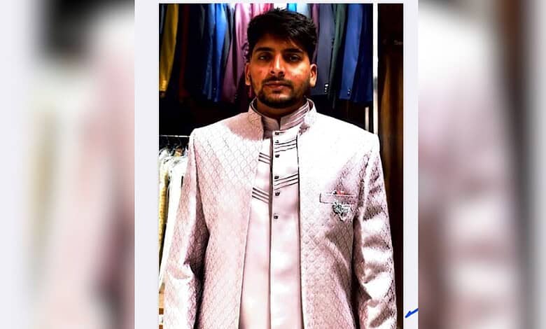Young man wearing a cream-coloured traditional outfit in a clothing shop with colourful garments behind.