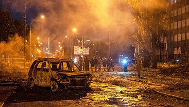 Burned-out vehicle and smoke in a city street after attack, with emergency responders present.