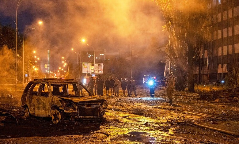 Burned-out vehicle and smoke in a city street after attack, with emergency responders present.