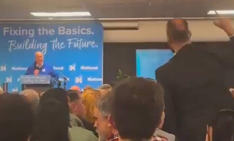 Protester confronts NZ PM Luxon during a political event with a crowd present.