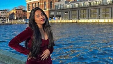 Young woman with long dark hair in a maroon dress by the river in an urban setting.