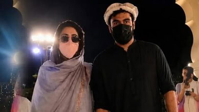 Two individuals wearing masks and traditional attire at an event with bright lights in the background.