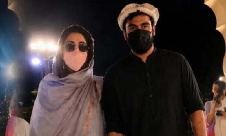 Two individuals wearing masks and traditional attire at an event with bright lights in the background.