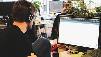 Young man with headphones working on a computer in a busy office environment.