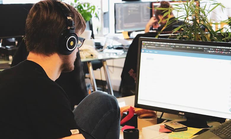 Young man with headphones working on a computer in a busy office environment.