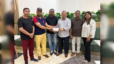 Group of six people in a room, one handing over a cheque to another, with plants in the background.