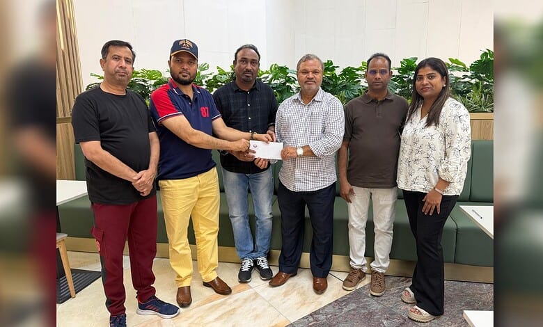 Group of six people in a room, one handing over a cheque to another, with plants in the background.
