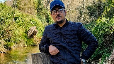 Man wearing glasses and a black jacket standing near a stream in nature.