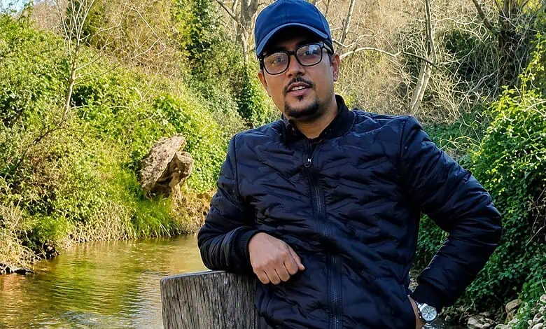 Man wearing glasses and a black jacket standing near a stream in nature.