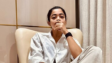 Rashmika Mandanna sitting on a cream sofa, wearing a white shirt, with a thoughtful expression.