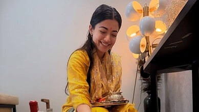 Rashmika Mandanna smiling while holding a plate of food in a cozy setting.