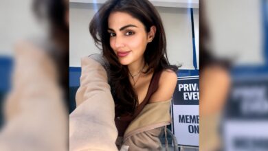 Rhea Chakraborty smiling in a casual setting with a beige jacket off her shoulder.