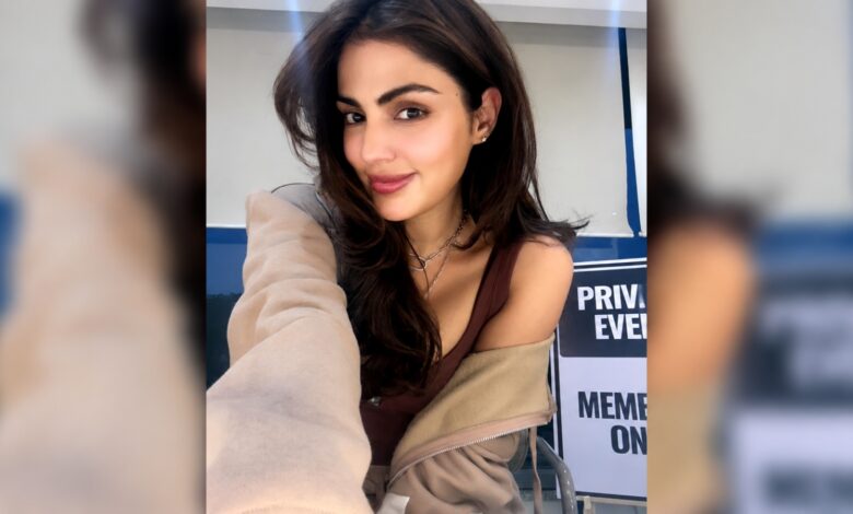 Rhea Chakraborty smiling in a casual setting with a beige jacket off her shoulder.