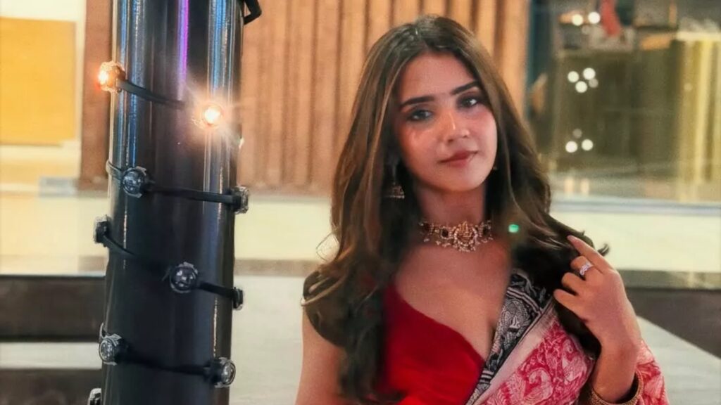 Young woman in red dress with long dark hair, wearing jewelry, in a well-lit indoor setting.