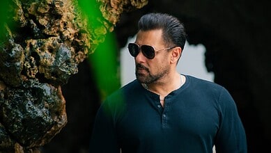 Salman Khan in a casual outfit with sunglasses outdoors near rocks and greenery.