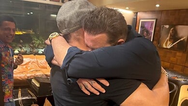 Two men hugging each other warmly in an indoor setting with a third man smiling in the background.