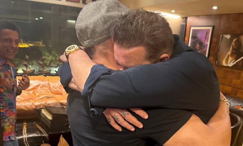 Two men hugging each other warmly in an indoor setting with a third man smiling in the background.